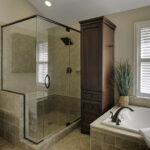 An elegant bathroom featuring a frameless glass shower enclosure; the elegant glass & shower mirrors reflect natural light from the shuttered windows onto earth-toned stone tiles.