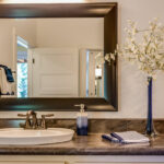 A large dark wood-framed mirror for the bathroom mounted above a marble countertop featuring a white vessel sink, blue decor accents, and a tall floral vase.
