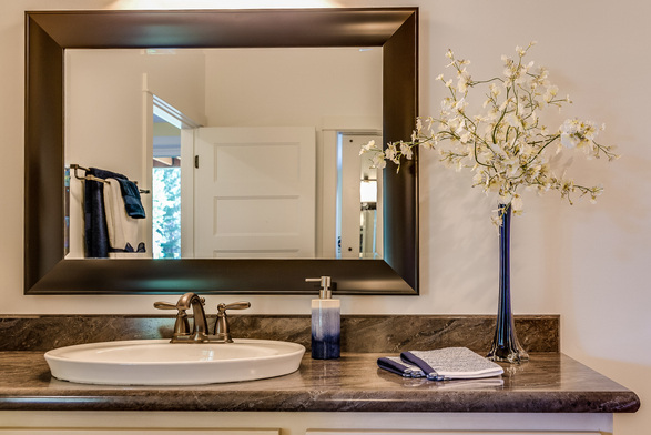 A large dark wood-framed mirror for the bathroom mounted above a marble countertop featuring a white vessel sink, blue decor accents, and a tall floral vase.